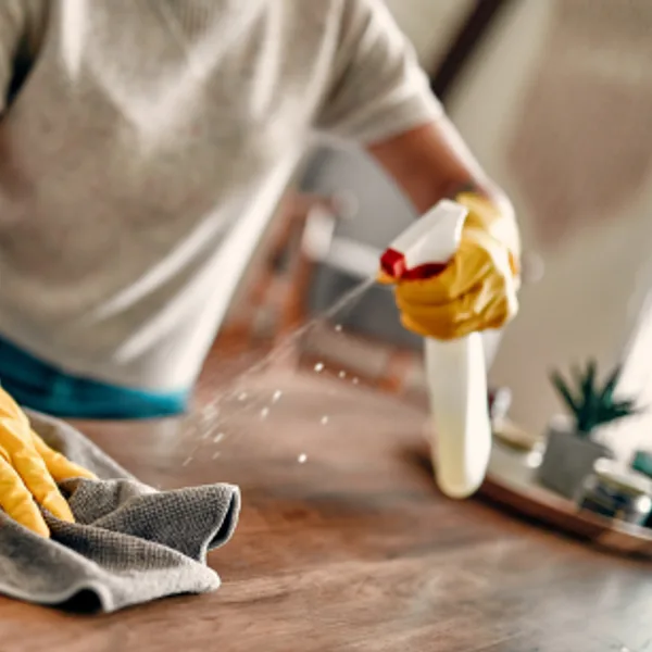 Gloved hand sanitizing a wooden table with a spray bottle and cloth